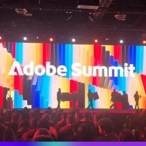Painel de palco iluminado com o logo do Adobe Summit em destaque, com uma tela de fundo colorida e várias pessoas no palco, em um evento de tecnologia.