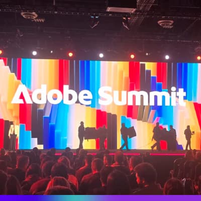 Painel de palco iluminado com o logo do Adobe Summit em destaque, com uma tela de fundo colorida e várias pessoas no palco, em um evento de tecnologia.