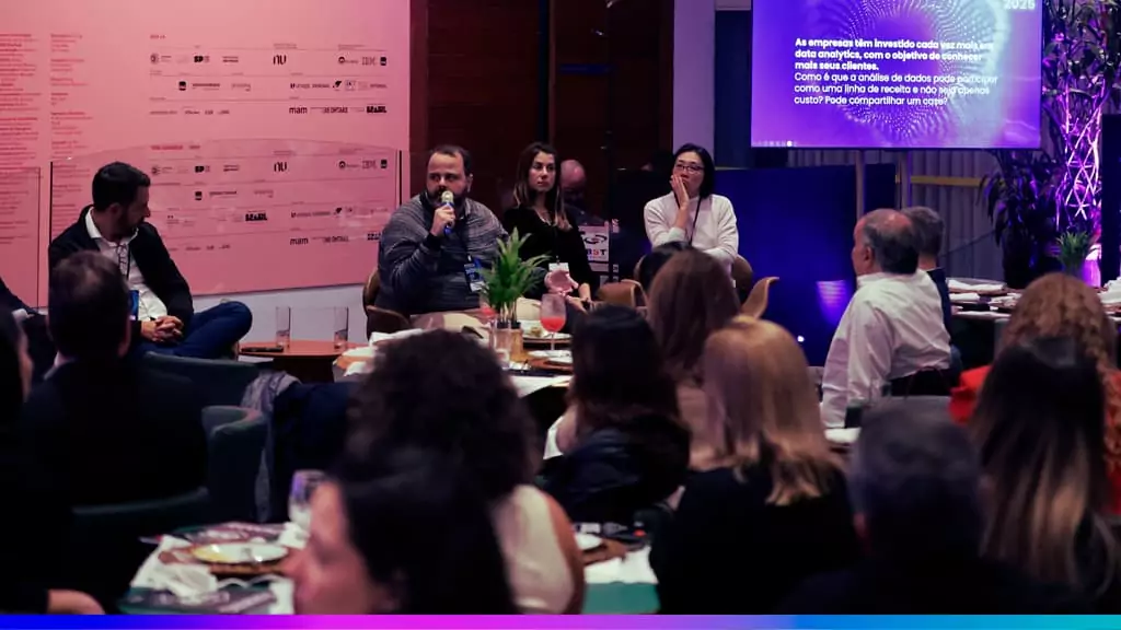Imagem de uma mesa redonda durante um evento de inova&ccedil;&atilde;o, com participantes discutindo, enquanto uma apresenta&ccedil;&atilde;o &eacute; exibida ao fundo.