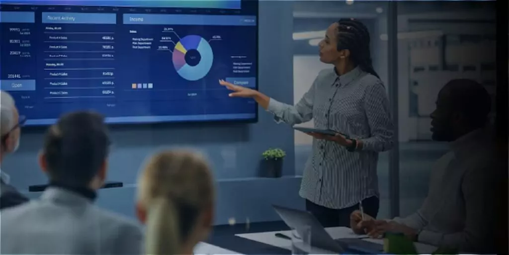 Mulher apresenta um dashboard de marketing digital em uma tela grande com gr&aacute;ficos e m&eacute;tricas de desempenho, enquanto uma equipe observa em uma sala de reuni&atilde;o.