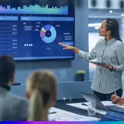 Mulher apresenta um dashboard de marketing digital em uma tela grande com gráficos e métricas de desempenho, enquanto uma equipe observa em uma sala de reunião.