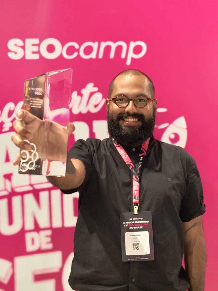 Analista de SEO Adonai Elias sorri enquanto segura o troféu no SEOcamp, como um dos vencedores do prêmio de melhor case de SEO do Brasil.
