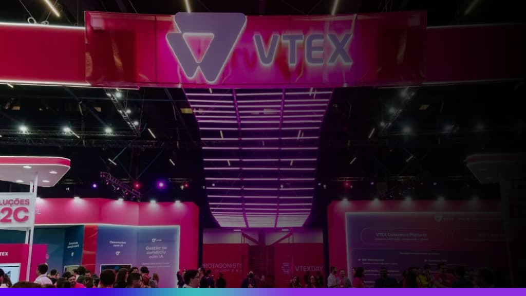 Estande da VTEX em evento, com estrutura iluminada em rosa e roxo, logo da marca em destaque no topo e público circulando pela feira.