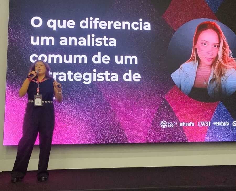 Jackeline Cavalcante, analista de SEO, apresenta uma palestra em um palco diante de um telão com o tema sobre a diferença entre um analista comum e um estrategista.