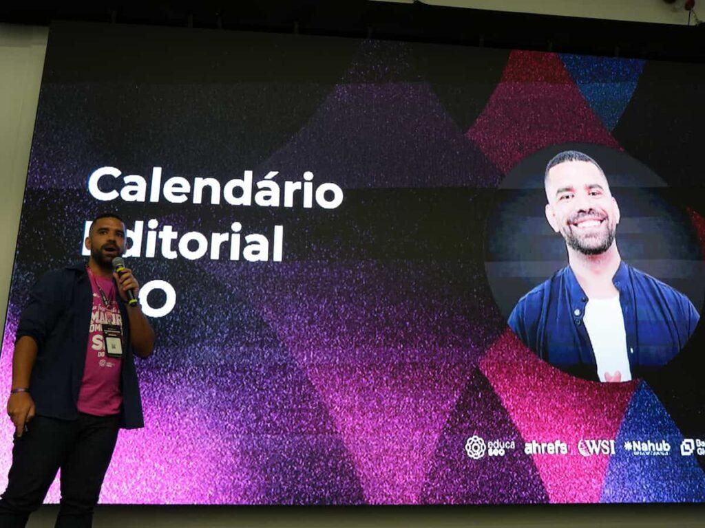 Luiz Paulo, analista de SEO, apresenta uma palestra em um palco, segurando um microfone diante de um telão com o tema “Calendário Editorial SEO”.